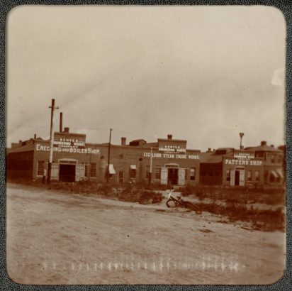 View of the Denver Engineering Works Company buildings on 30th (Thirtieth) and Blake Streets in Denver, Colorado. A bicycle lies near the dirt street. The brick buildings read: "Erec[t]ing And Boiler Shop", "Exc[e]lsior Steam Engine Works", and "Pattern Shop".