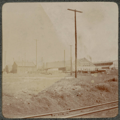 View of the Colorado Iron Works shops on 33rd (Thirty-third) and Wynkoop Streets in Denver, Colorado. Metal tubes are outside the buildings that read: "Mining Machinery" and "Car Wheels & Gagings". Union Pacific Railway Company (UP) tracks are nearby.