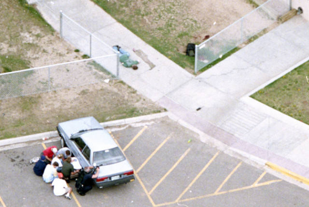 A student lies on a sidewalk while an officer guards others behind a car.