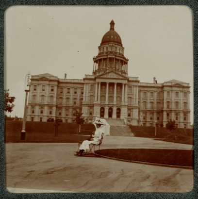 A woman poses with a parasol on a bench in Civic Center Park near the Colorado State Capitol building in Denver, Colorado. She wears a long dress and hat.