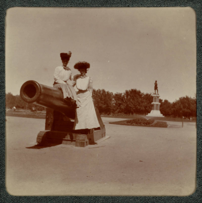 Women in long dresses and hats pose on a Naval artillery (a rifled Parrott) cannon in City Park, Denver, Colorado. A statue of Robert Burns is in the distance.