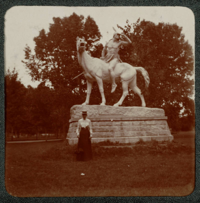 A woman poses near "The Indian," a sculpture by Alexander Phimister (A. P.) Proctor, in City Park, Denver, Colorado. The sculpture depicts a Native American man who holds a spear on horseback.