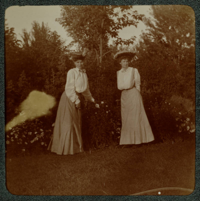 Women pose near flower bushes in a garden probably in Denver, Colorado. They wear long dresses and hats.