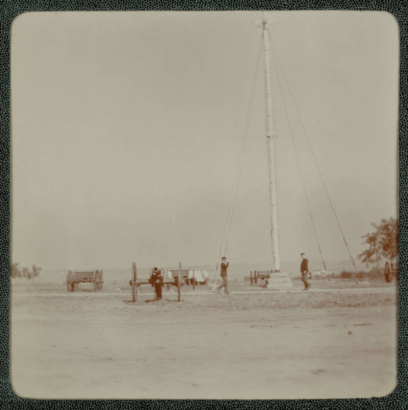 Men in military uniforms walk near a flagpole possibly in Denver, Colorado. Cannons and wooden wagons are nearby. The flagpole is tethered by guy wires.