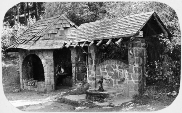 View of the spring house at the upper switch back of the Lariat Loop Road on the way to Lookout Mountain Park, a Denver Mountain Park located 17 miles west of Denver, Colorado. Shows two rock ashlar buildings and a water pump in front of a arched rock wall. The shelter has an arch window, a stone bench and shingled roof.
