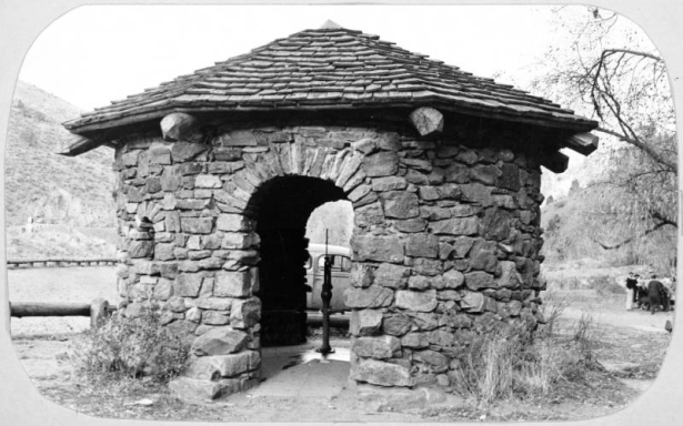 A view of a pump house at Little Park, Jefferson County, located 21 miles southwest of Denver, Colorado. The round shelter has rock ashlar walls, two arched doorways and a shingled pyramidal roof. The pump is in the center of the shelter.