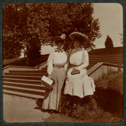 Women pose near a set of stairs, probably in City Park, Denver, Colorado. They hold handbags and a box and wear long dresses and hats.
