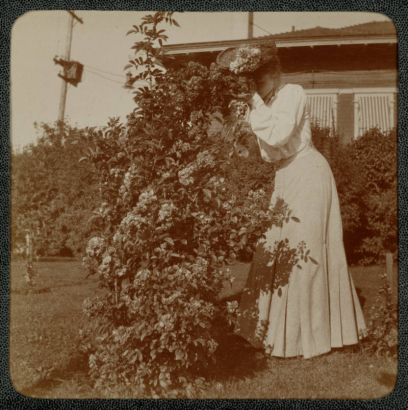 A woman poses outdoors near a shrub at her home in Denver, Colorado. She wears a long dress and a hat decorated with flowers.