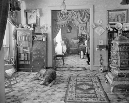 Interior view of the Finding residence in Breckenridge (Summit County), Colorado; decor includes china plates, wicker chairs, a pitcher,a table made of machine parts, books, a seashell, a stove, drapes, a combination bookcase, plants, fans, ingrain carpet, oriental rugs, cut paper dainties, and greeting cards. A dog sleeps on the floor.