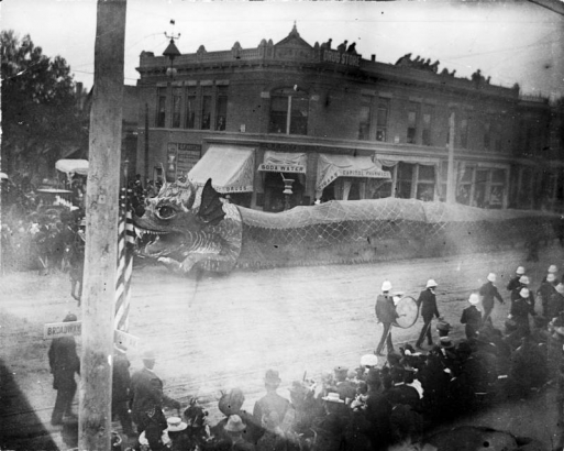 View of a Chinese dragon and marching band on Broadway near 14th (Fourteenth) Street in Denver, Colorado. The parade is part of the Festival of Mountain and Plain. Spectators line the street. Signs on nearby buildings read: "G. F. Hatch," "Soda Water," "Cigars," "Drug Store," and "Capitol Pharmacy."