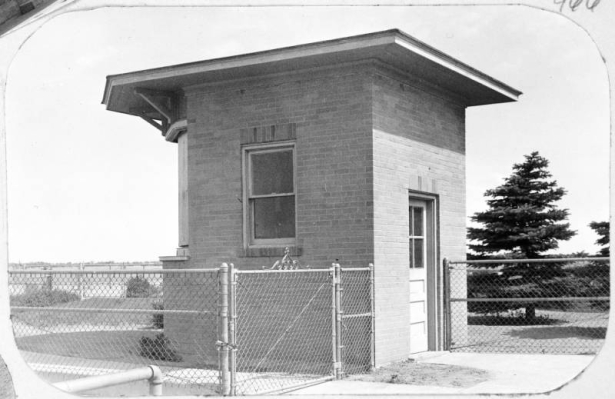 Shows the small brick lighting control building at the Denver Municipal Airport (later Stapleton International Airport) in Denver, Colorado. The building features a single door, windows and a flat roof with an overhang; a chain link fence is in front of the building.