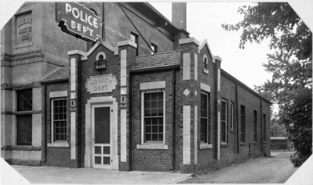 An exterior view of Police Station number 4 located on West 26th Avenue and Federal Boulevard in Denver, Colorado. The single story brick building features cement trim and accents and a shingle roof. A sign above the entrance reads: "Police Dept."; and a neon sign above the building reads: "Police Dep't".