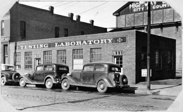 Automobiles are parked in front of the single story brick testing laboratory of the Denver Highway Department located near 8th (eighth) and Walnut Street in Denver, Colorado. Address above two doors of the building read: "811" and "805". Garage number one is in the background.