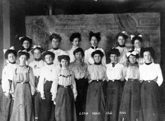 Sixteen members of the Leap Year Club in Basalt, Colorado pose in  two lines. Ten women are in the first row, and six women are in the second row. Characteristic of the early twentieth century, their clothing includes long-sleeved, high-collar blouses with puffed sleeves and long skirts. Several of the women wear jewelry, including earrings, cameo broaches, and pendants. A painting of an ornate room is painted on the wall behind them.