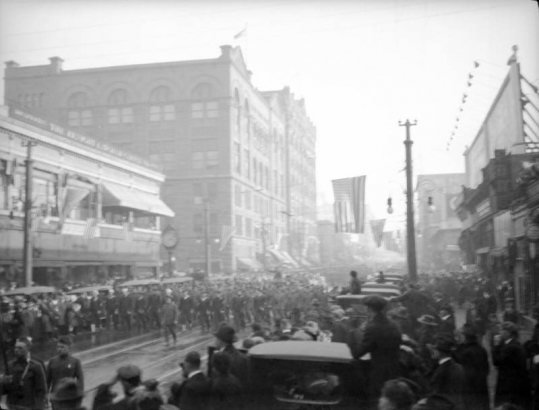 Men and women march in a parade to commemorate the end of World War I, on 16th (Sixteenth) Street in Denver, Colorado. Spectators line the streets and U. S. flags fly from nearby buildings.