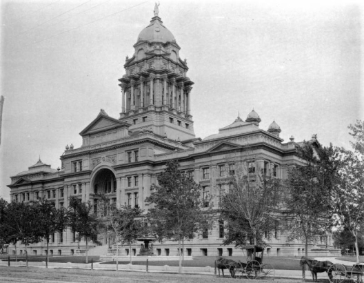 Exterior view of the Arapahoe (later Denver) County Court House, designed by architect Elijah E. Meyers, located between 15th, 16th and Tremont Streets and Court Place in Denver, Colorado. Shows horse-drawn carriages and a fountain on the lawn of the county building.