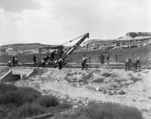 Denver and Rio Grande Western Railroad construction in Ruby Canyon, (probably) Utah consists of men laying track over a concrete bridge, using a rail-adapted crane. Sandstone cliffs are in the background.