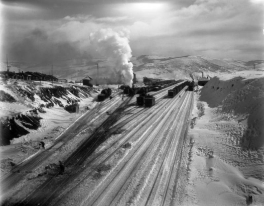 Soldier Summit, Utah, is a Denver and Rio Grande Western Railroad terminal/switch yard covered in snow. Locomotives, freight and passenger cars, and cabooses are on the tracks, and men work picking ice from the switches. Passengers emerge at the depot; employee houses dot the hill to the side.