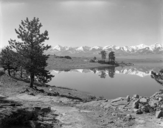 Trees and mountains are reflected in De Weese Reservoir, Custer County, Colorado; wide fields and the Sangre De Cristo Range are in the background.