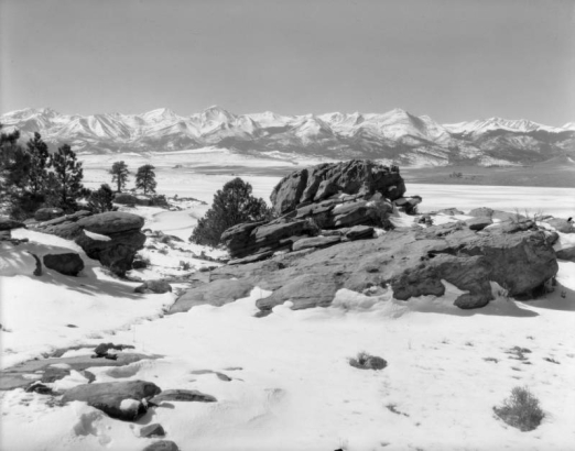 Rocks, trees, and wide fields edge De Weese Reservoir in Custer County, Colorado; the Sangre De Cristo Range is in the distance.