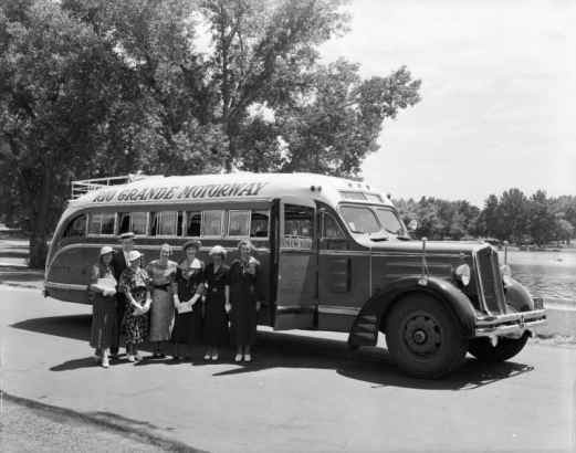 View of a Rio Grande Motor Way passenger bus. Women and a man pose by the 1935 White brand vehicle; outfits include print dresses and straw hats. Pin striping reads: "Los Angeles - Salt Lake - Grand Junction - Pueblo - Denver - Omaha - Kansas City - Chicago - New York 154;" possibly City Park Lake is in the background.