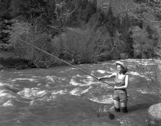 A young woman fishes with a bamboo pole, in (probably) Colorado or Utah; she wears hip waders and stands in the river. Her outfit also includes short-legged bib-overalls and a straw hat.