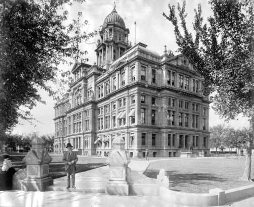 View of the Denver (originally Arapahoe) County Courthouse (demolished in 1933) located in the block of 15th (Fifteenth) and 16th (Sixteenth) Streets and Tremont and Court Places in Denver, Colorado. The Greek Revival building has classical entablature and a dome. Sign reads: "W. F. Hynes, Justice of the Peace." Bicycles lean against a fence.