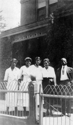 African American women who include Fairfax Holmes pose with a Black man near the Phyllis Wheatley Branch YWCA at 2460 Welton Street in the Five Points neighborhood of Denver, Colorado. They stand near a metal wire fence.
