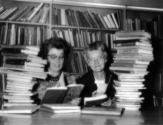 Two librarians from the Aurora Public Library in Aurora, Colorado, sit at a table, partially hidden by piles of books. Behind them are bookshelves full of more books. They each hold open books and book cards in their hands.