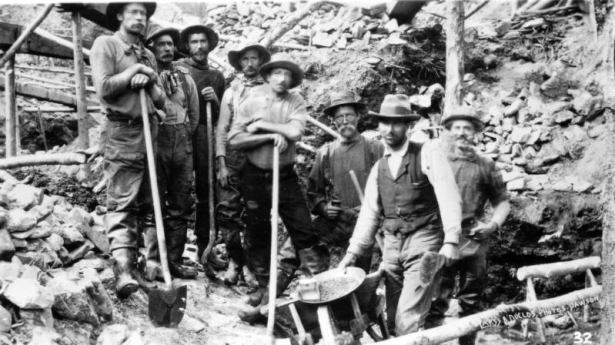 A group of miners pose standing near a wheelbarrow, Yukon Territory. One man holds a pan full of placer ore, the others hold shovels; mining construction is behind them.