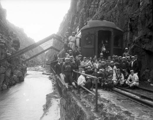 Men, women, children, and the Chicago White Sox pose by a Denver and Rio Grande passenger car and the Arkansas River at Hanging Bridge, in the Royal Gorge, Fremont County, Colorado. Baseball team owner Charles A. Comiskey wears a fedora hat.