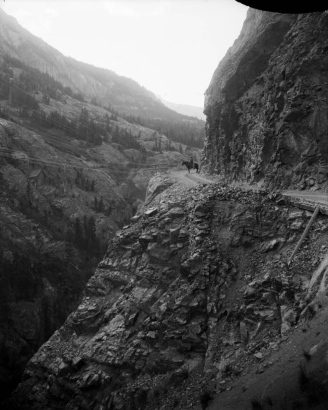 View of the Otto Mears toll road in Uncompahgre Gorge, Ouray County, Colorado; shows a man on a horse, power poles, and cliffs.