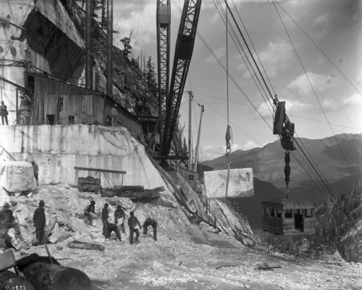 View of the Colorado Yule Marble Company quarry, four miles south of Marble (Gunnison County), Colorado; shows a crane, buildings, cable and pulleys, a suspended block of stone, a gondola, and men working. The block will be transferred to the cable the gondola is on for lowering to the standard gauge electric railway for transport to the mill site, in the town of Marble.