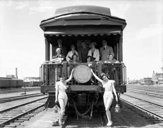 Men and women pose on the observation platform of the "Glenwood Canon" Denver and Rio Grande Western Railroad passenger coach at Burnham Shops, Denver, Colorado. Ladies in bathing suits and conductor's hats pose with lanterns.