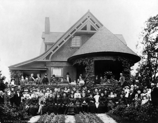 Portrait of well-dressed men, women, and children at a house in Denver, Colorado; costume includes bodices, flounces, straw, bowler, and stovepipe hats. The residence has shingle imbrication, a round terrace, and bargeboard.