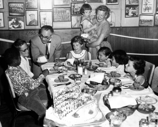 Interior view of the Ringside Lounge at 1120 17th Street in Denver, Colorado; shows boys, girls, a man and a woman at a table set with food and dinnerware. Joe "Awful" Coffee offers a piece of cake; Jane Sterling holds a child, and framed photographs cover the wall.