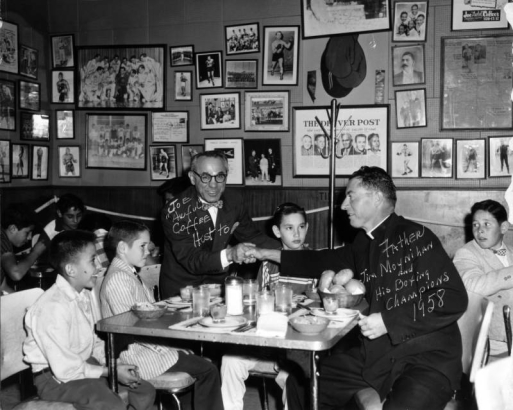 Interior view of the Ringside Lounge in Denver, Colorado; shows boy boxers, Joe "Awful" Coffee shaking hands with Father Jim Moynihan, and a table set with food. Framed pictures of boxers and newspaper clippings cover the wall.