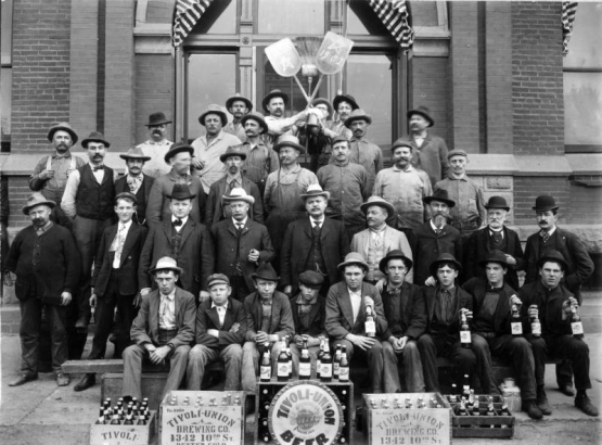 Men and boys pose with cigars, pipes, shovels, a broom, and beer bottles. Crates and labels read: "Tivoli Union Brewing Co. 1342 10th St Denver, Colo."