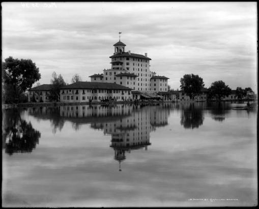 Exterior, back view of the Broadmoor Hotel in Colorado Springs, Colorado; surface of lake reflects hotel image; men and horse-drawn wagon are on opposite shore.