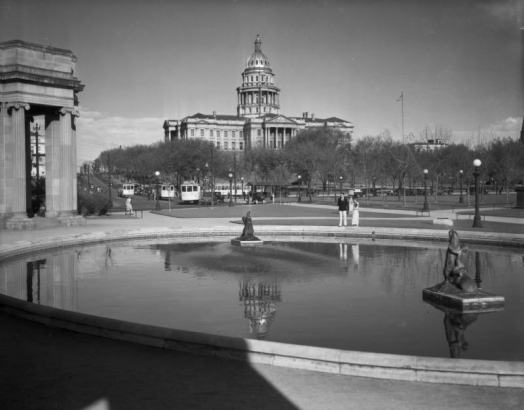 View of the Colorado State Capitol building, Denver, Colorado; shows people standing by the Civic Center reflecting pool and colonnade, trees, and streetcars of the Denver Tramway Company.