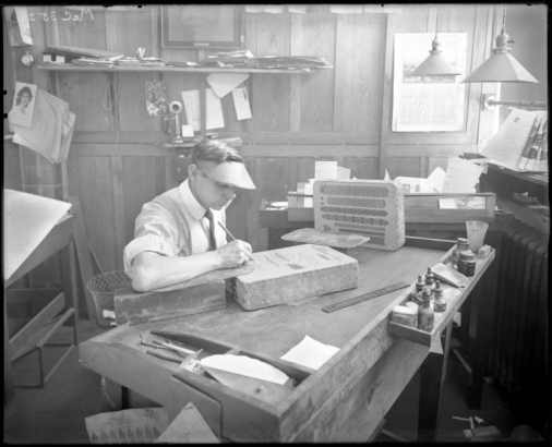 A printer sits at a table, working with printing blocks at the Smith-Brooks Printing Company in Denver, Colorado; shows ink bottles, company name, and July 1914 calendar posted on wall.