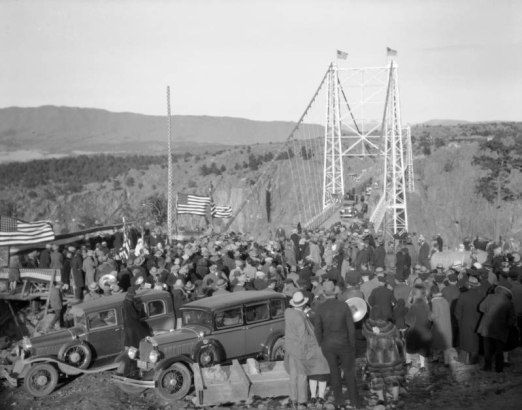 People crowd the Royal Gorge Bridge for its dedication in Fremont County, Colorado. A woman in a fur coat is by automobiles; United States flags and bunting decorate a dais.