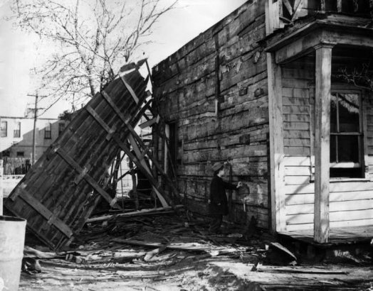 A boy stands in ruins of a frame building identified as the first hotel in Denver, Colorado; he looks at a hewn log wall.