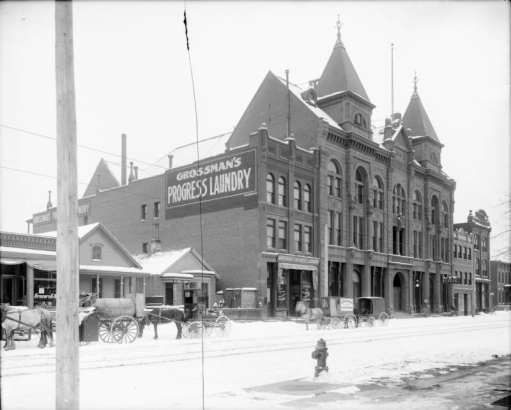 Horse-drawn delivery wagons are parked near the Turnhalle and other buildings at 22nd (Twenty-second) and Arapahoe Streets in Denver, Colorado. Signs on the wagons read: "Texas Oil Company Petroleum and Its Products" and "Grossman's Progress Laundry."