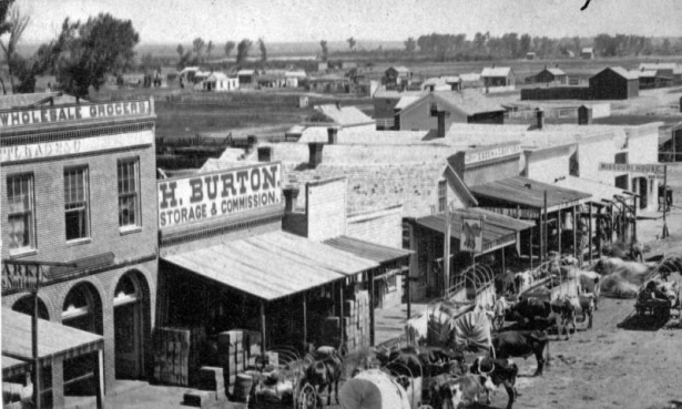 Horse-drawn wagons are parked near buildings on Blake Street in downtown Denver, Colorado. Signs on the buildings read: "Wholesale Groceries," "H. Burton Storage & Commission," and "Missouri House." The hotel has other signs: "Bar Room," "Boarding," and long horned cattle are tethered among the horses. Water of the South Platte River is in the background.