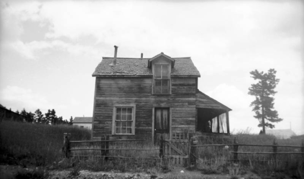 View of a large, dilapidated house in Gold Hill, Colorado; features a side gable, gabled dormer, an attached, closed porch, single leaf door, double hung windows, and a horizontal timber fence with a picket gate that surrounds the property; tall grass in the yard and surrounding area.