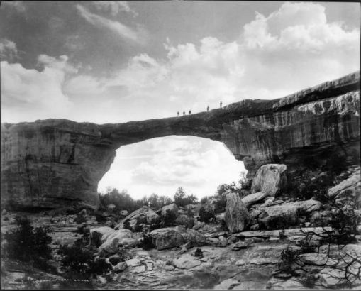 Owachomo Natural Bridge, Armstrong Canyon, Natural Bridges National Monument, San Juan County, near Blanding, Utah. People pose on top of the sandstone formation also known as Little Bridge, with pinyon and  juniper trees on slope below.