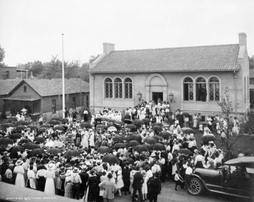 People, holding umbrellas or United States flags, crowd the opening dedication ceremonies for the Byers Branch of the Denver Public Library, 675 Santa Fe Drive, Denver, Colorado. Boys climb on an automobile by the  stucco - brick building with tile roof, arched windows and entrance.