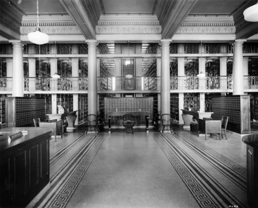 Interior of the (Carnegie) Main Public Library, Civic Center, Denver, Colorado; features columns, ornate stenciled beams with moldings and dentils separating ceiling bays and a geometric pattern tile floor. The circulation desk is by catalog cabinets, bookshelves, chairs and wood tables; signs read: "Information," and "Return Desk."