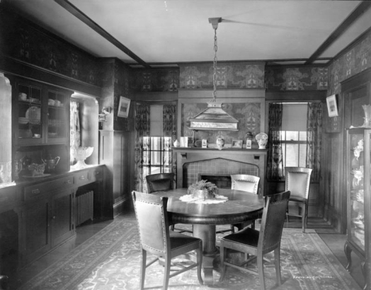 Interior view of a Denver, Colorado, residence; dining room furnishings include a brick fireplace, wood paneling, molding, Art Nouveau wallpaper, a pedestal table, leather upholstered wood chairs, Oriental rug,  stained glass lamp, built-in buffet, crystal punch bowl, silver coffee pot, and china cabinet. Vases and photographs top the mantle.